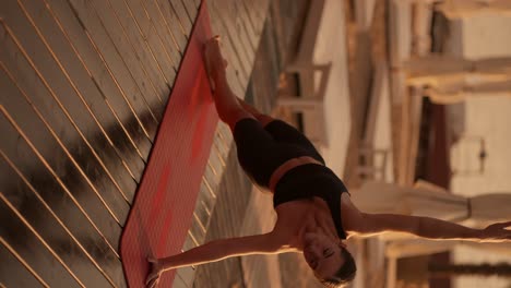 vertical video: a brunette girl in a black sports summer uniform does a plank on one arm and stretches up with the other on a sunny beach covered with boards. yoga and stretching classes during golden sunset on the beach in summer