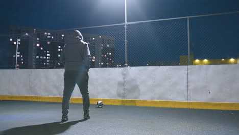 man in hoodie kicking soccer ball against wall on illuminated urban sports field at night, with reflection visible on wall and city lights glowing in background