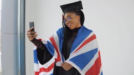 happy stylish afro american female student in graduate uniform taking selfie holding british flag on shoulders