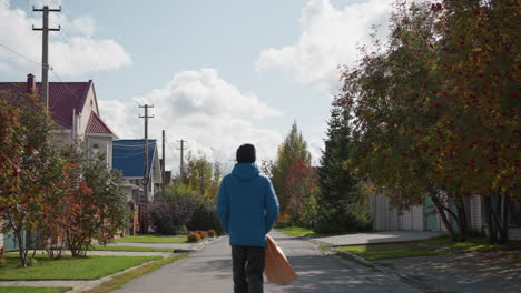 boy in blue hoodie and black beanie walks down residential street with paper bag, surrounded by autumn foliage, and colorful houses under cloudy skies