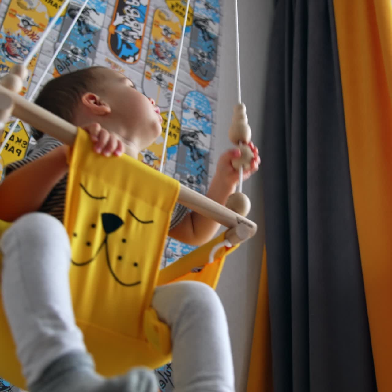 Lovely little baby swinging in the swing in the room. Kid plays with beads on the rope of a swing. Low angle view