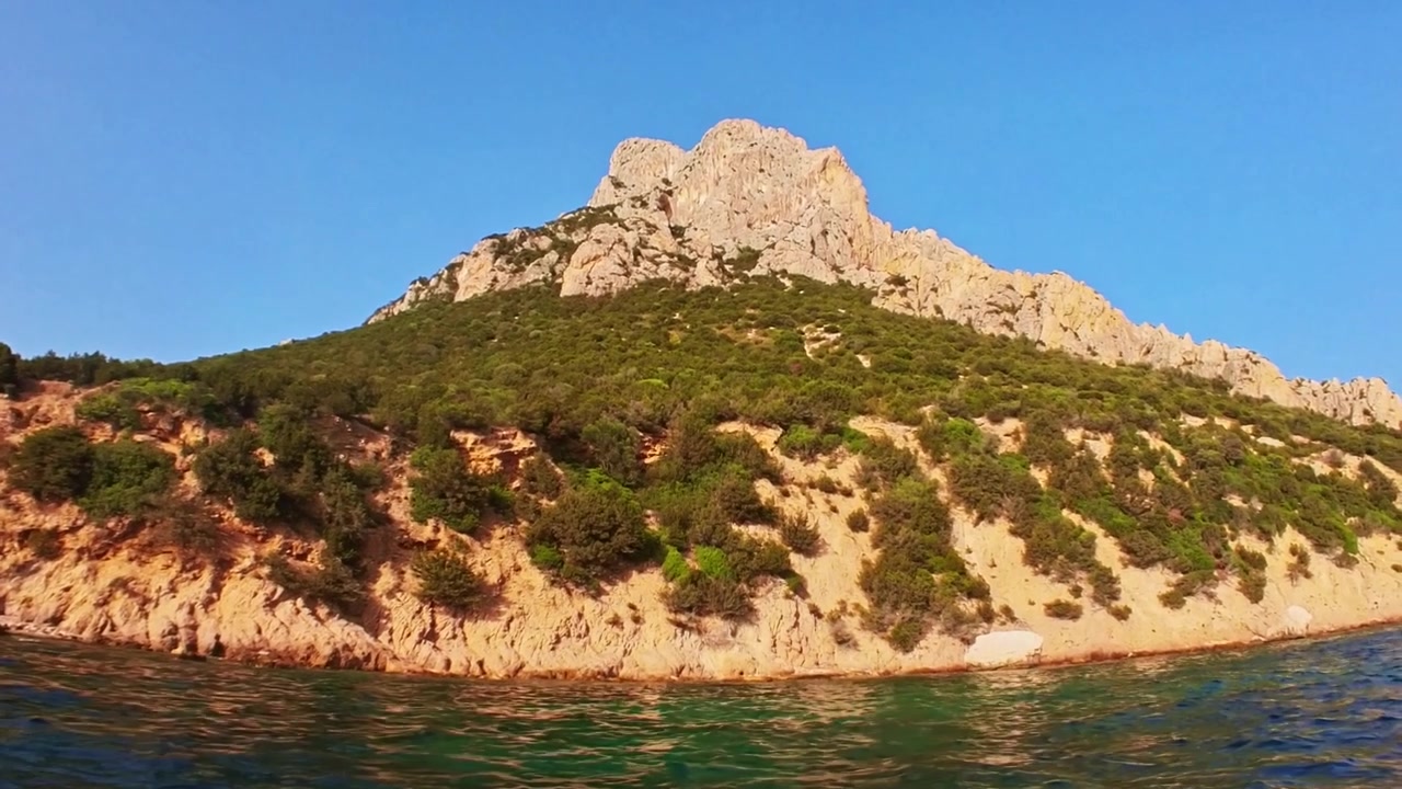Punto di vista da una barca che naviga lungo una costa frastagliata con montagne rocciose, lussureggiante vegetazione mediterranea e acqua limpida color smeraldo