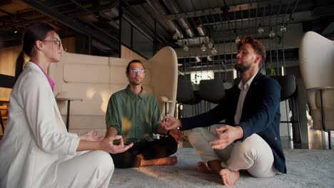 3 office workers, a girl in a white suit, a guy in a green shirt and a guy in a blue jacket are doing meditation on a mat in the office. meditation and zen practice at work