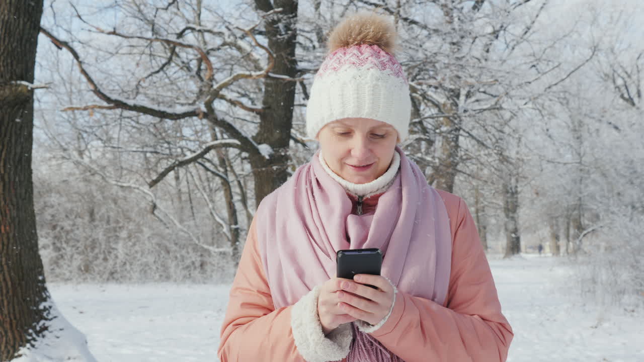 분홍색 재킷을 입은 한 여성이 겨울 공원에서 산책을 즐깁니다. 휴대전화 4k 슬로우모션 비디오를 사용합니다.