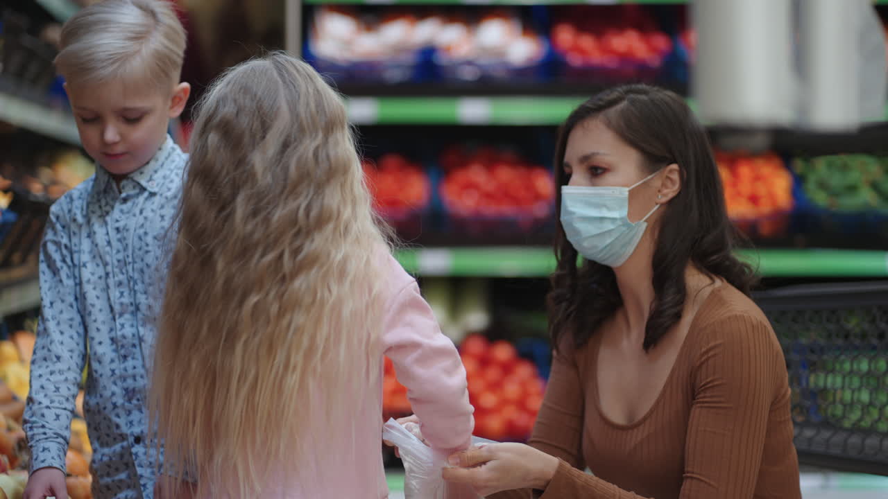 una madre con una máscara protectora con dos hijos está comprando comestibles en el supermercado. comprando alimentos, verduras y frutas con niños