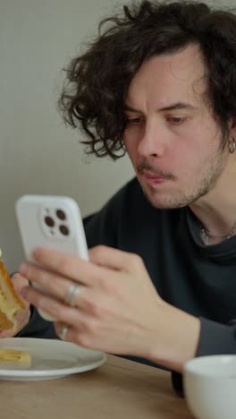 vertical video of a brunette guy with a mustache with curly hair sitting at a table and watching videos on social networks while eating his sandwich in the morning