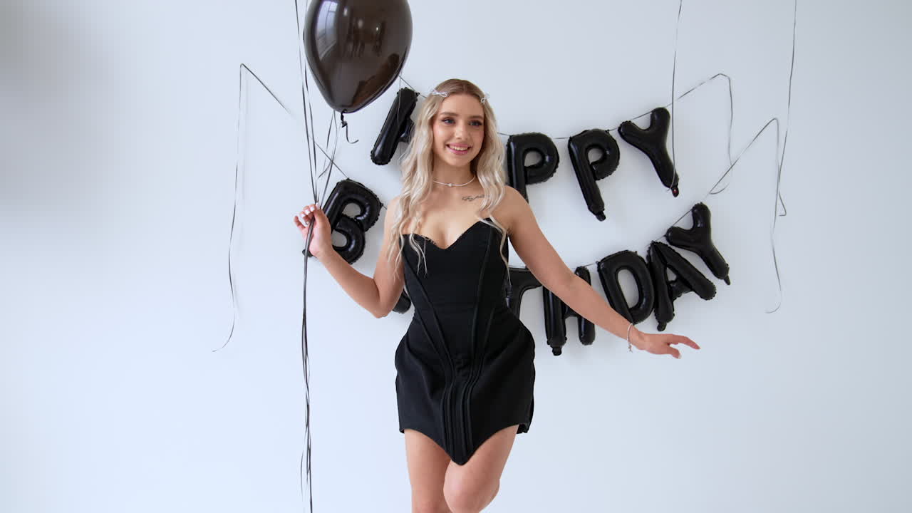 Blonde Caucasian woman in short black dress holds a bunch of balloons. Black Happy Birthday sign on the wall at backdrop.
