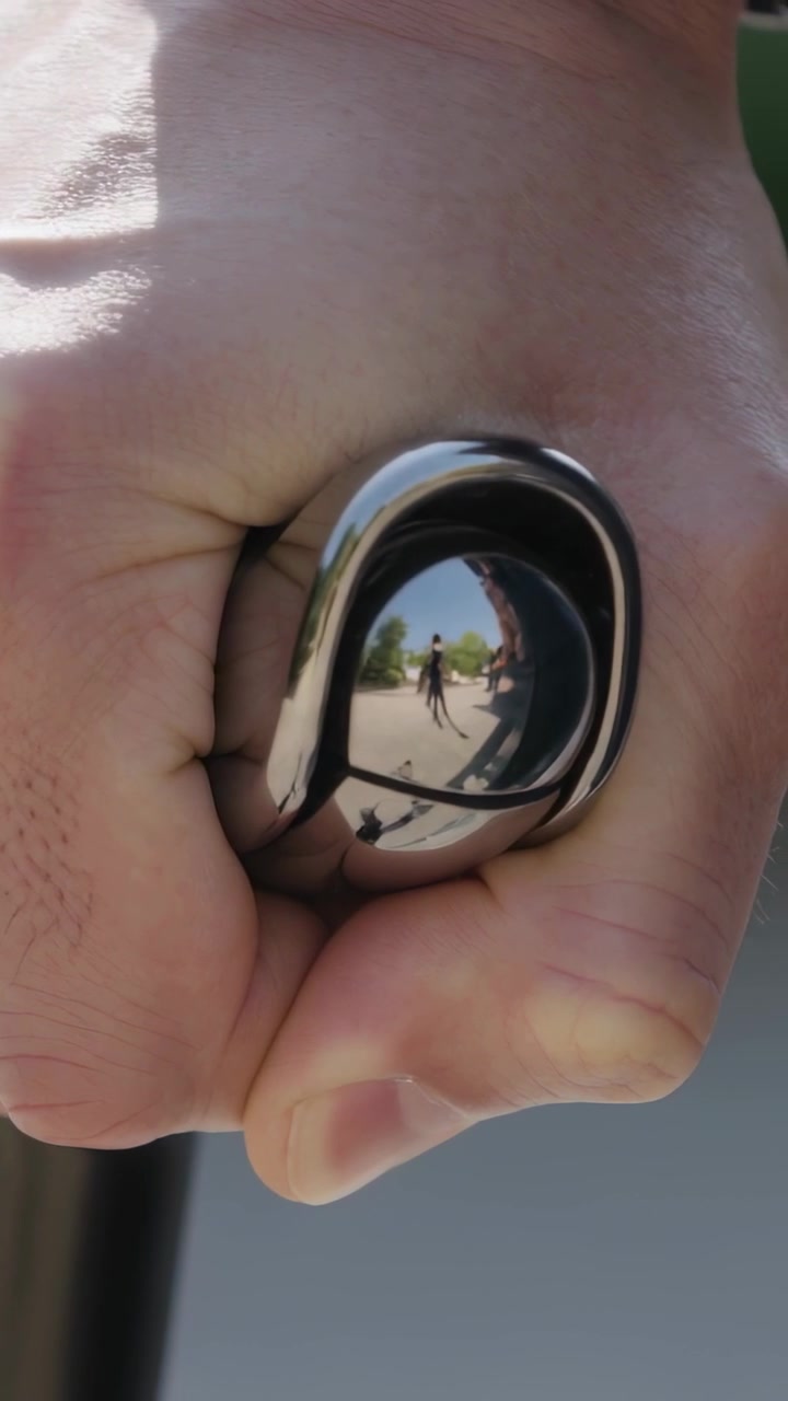 Video verticale: Mano destra che inclina e afferra un oggetto lucido a forma di goccia sul marciapiede, rivelando una fotocamera e una persona.