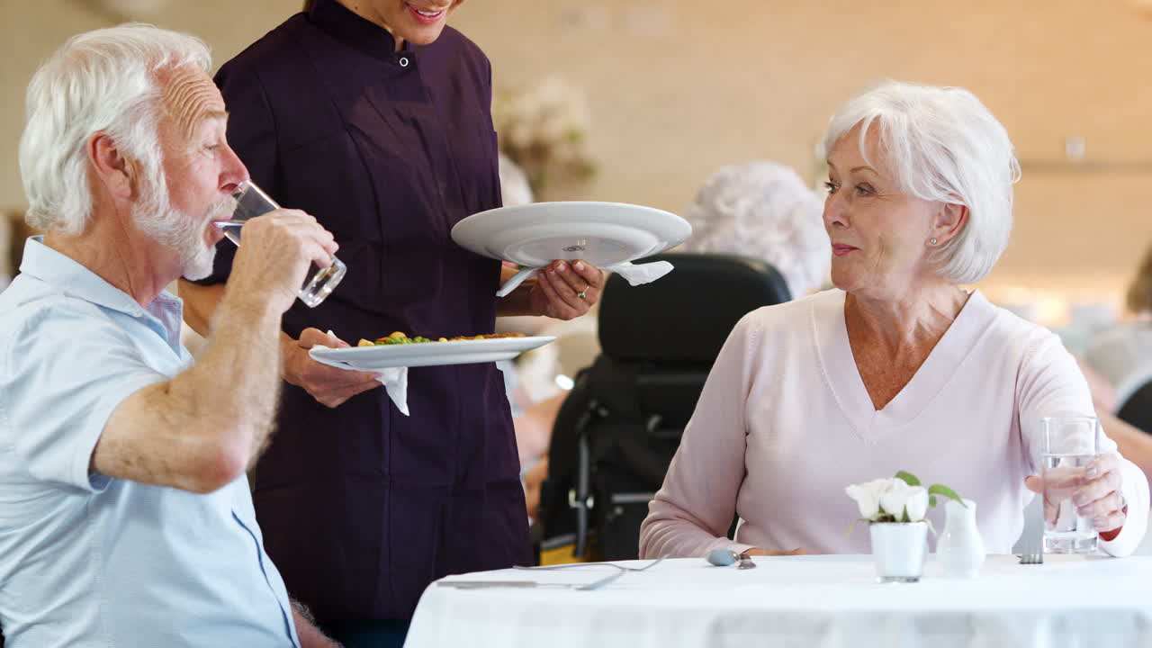 pareja de ancianos siendo servida con una comida por un cuidador en el comedor de un hogar de ancianos