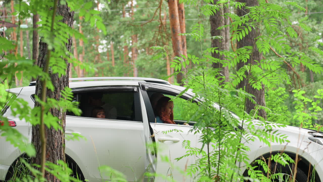 Young woman with colored hair wearing long robe enters parked car beside husband and child in lush green arboretum, soft daylight filtering through leaves, serene family moment in nature setting
