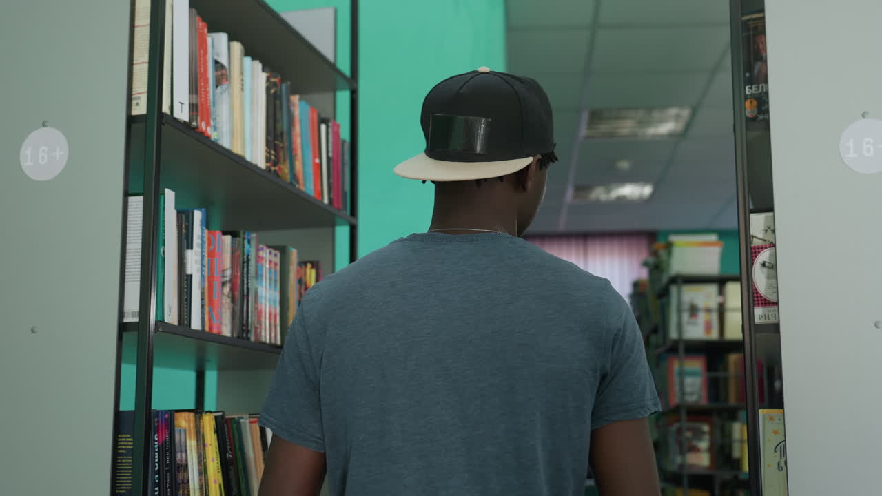 young man wearing casual outfit with cap backwards sits cross egged in library aisle reading open book surrounded by tall bookshelves filled with book