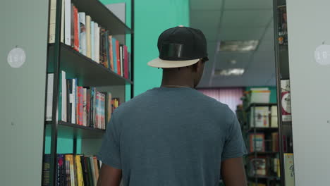 young man wearing casual outfit with cap backwards sits cross egged in library aisle reading open book surrounded by tall bookshelves filled with book