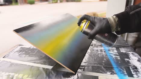 Man-with-black-gloves-holding-spray-paint-can-spraying-it-across-a-wooden-canvas-board-with-blue-yellow-black-art-on-street-in-background