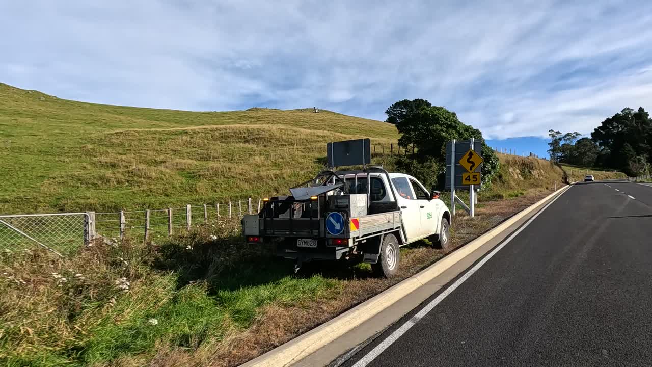 A truck and worker conduct road maintenance on a rural road in Akaroa, New Zealand, under clear skies and natural lighting