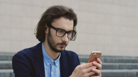 Retrato-Del-Joven-Hombre-De-Negocios-Triste-Con-Anteojos-Que-Parece-Preocupado-Mientras-Envía-Mensajes-De-Texto-Y-Chatea-En-El-Teléfono-Inteligente