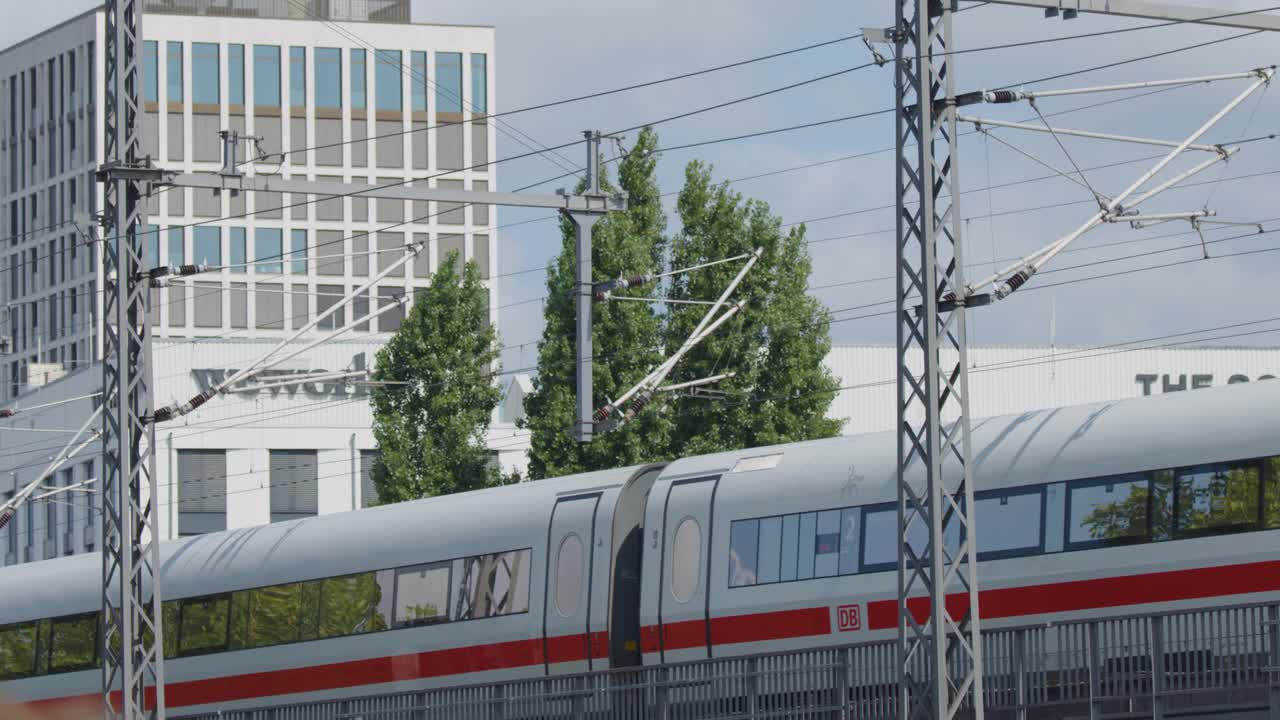 A modern white high-speed passenger train with graffiti moves quickly past urban buildings and power lines in Berlin, Germany, under bright daylight with steady camera tracking