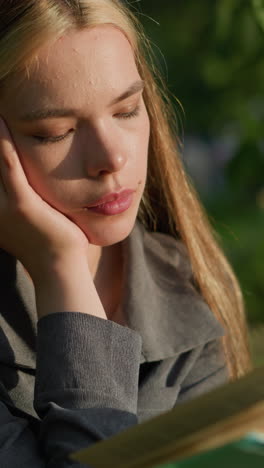 lady in grey clothing seated under tree, head resting on left hand while reading book, looking moody, sunlight softly illuminates her face, with background slightly blurred, featuring greenery