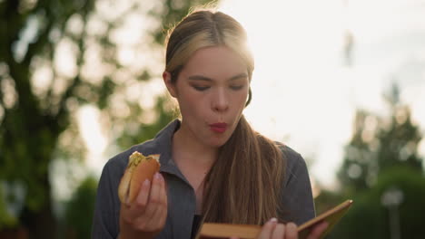 dama de camiseta gris leyendo un libro mientras sostiene una hamburguesa, masticando pensativamente con la suave luz del sol iluminando la escena, el fondo borroso por los rayos del sol y la vegetación