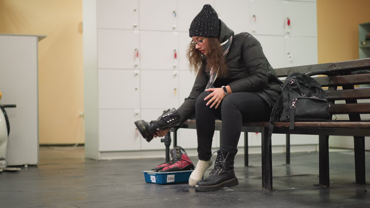 Female sitting on wooden bench in locker room wearing black coat and knitted hat lacing black boots next to blue tray with red skates preparing for winter skating activity with backpack on bench background
