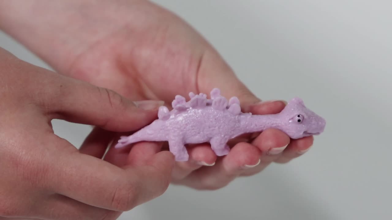 Händer som sträcker och klämmer en lila dinosaurieleksak av gummi mot en vit bakgrund