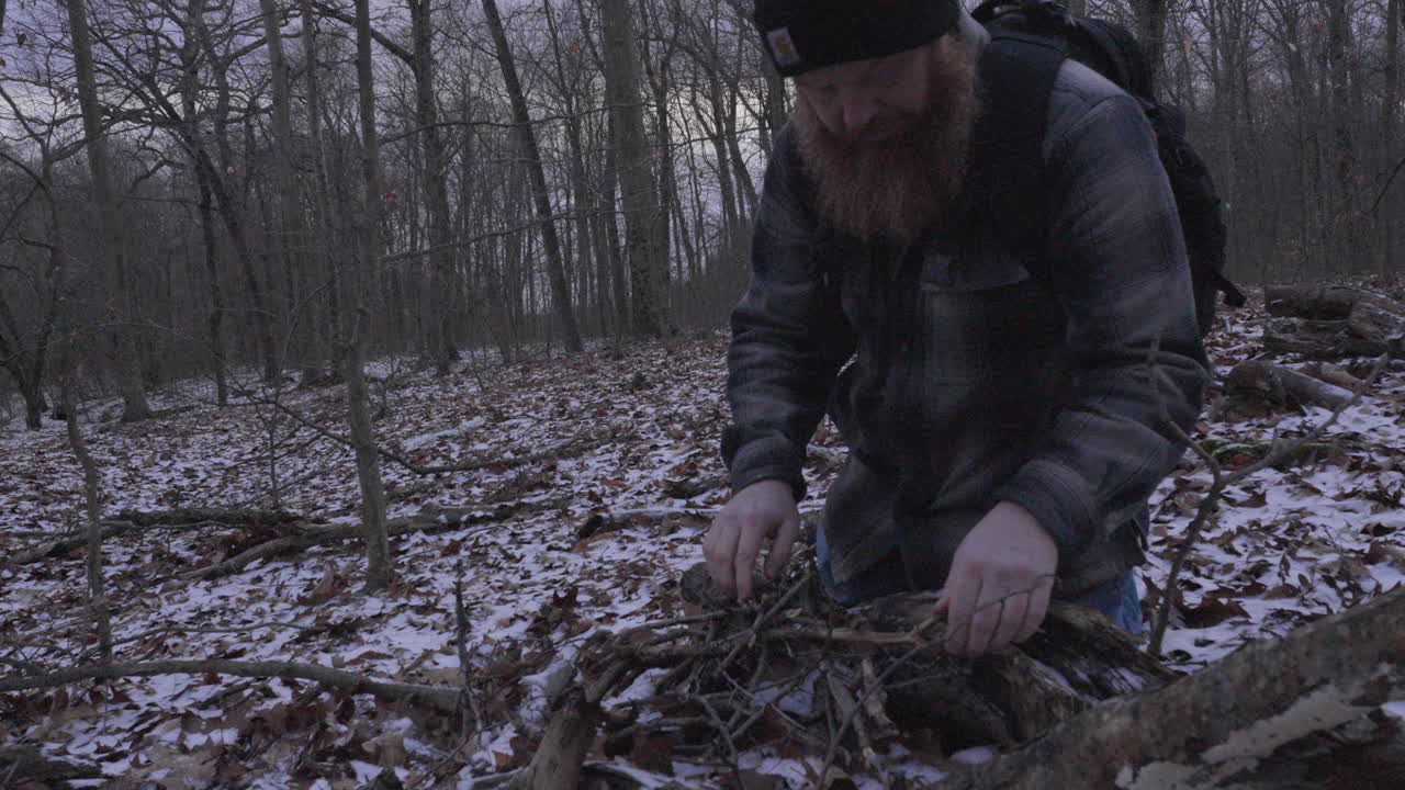 movimiento lento de un hombre con una gran barba roja y con una camisa de franela a cuadros recoge leña rompiéndola en leña y apilándola para una fogata en un entorno forestal con nieve en el suelo