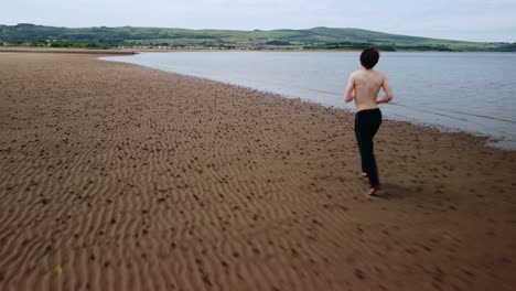 A-young-man-running-on-a-beach-with-no-shirt-or-shoes-on,-aerial