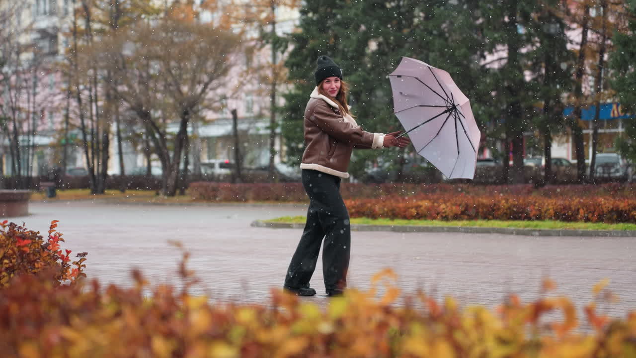 Cheerful girl holding umbrella, wearing knit cap, brown shearling jacket, black trousers, happily turning around in light snowfall, enjoying autumn day, embracing cold weather with joy