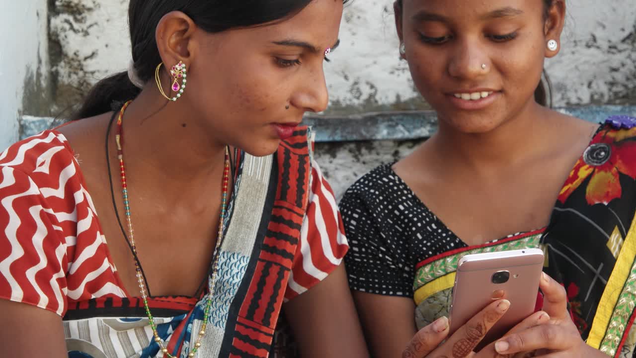 전통적인 인도 의상을 입은 두 명의 아름다운 여성이 휴대 전화로 집의 편안함에서 서로 사진 비디오를 공유합니다.