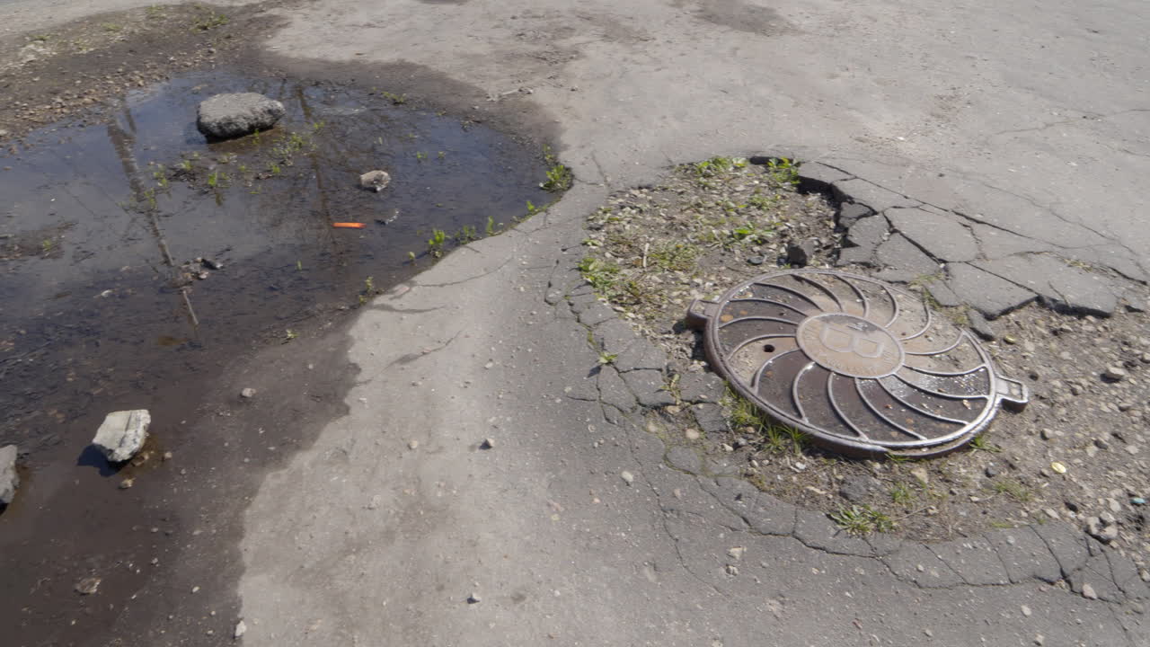 Broken drain hatch with cracks in asphalt pit roads pedestrian walkway, panning shot