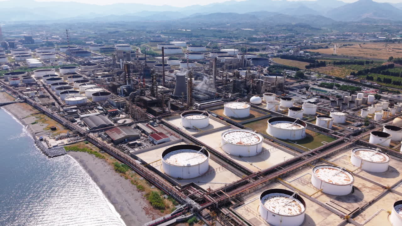 Drone flies high above an oil refinery in Sicily, showing massive tanks, industrial structures, shoreline, and distant mountains