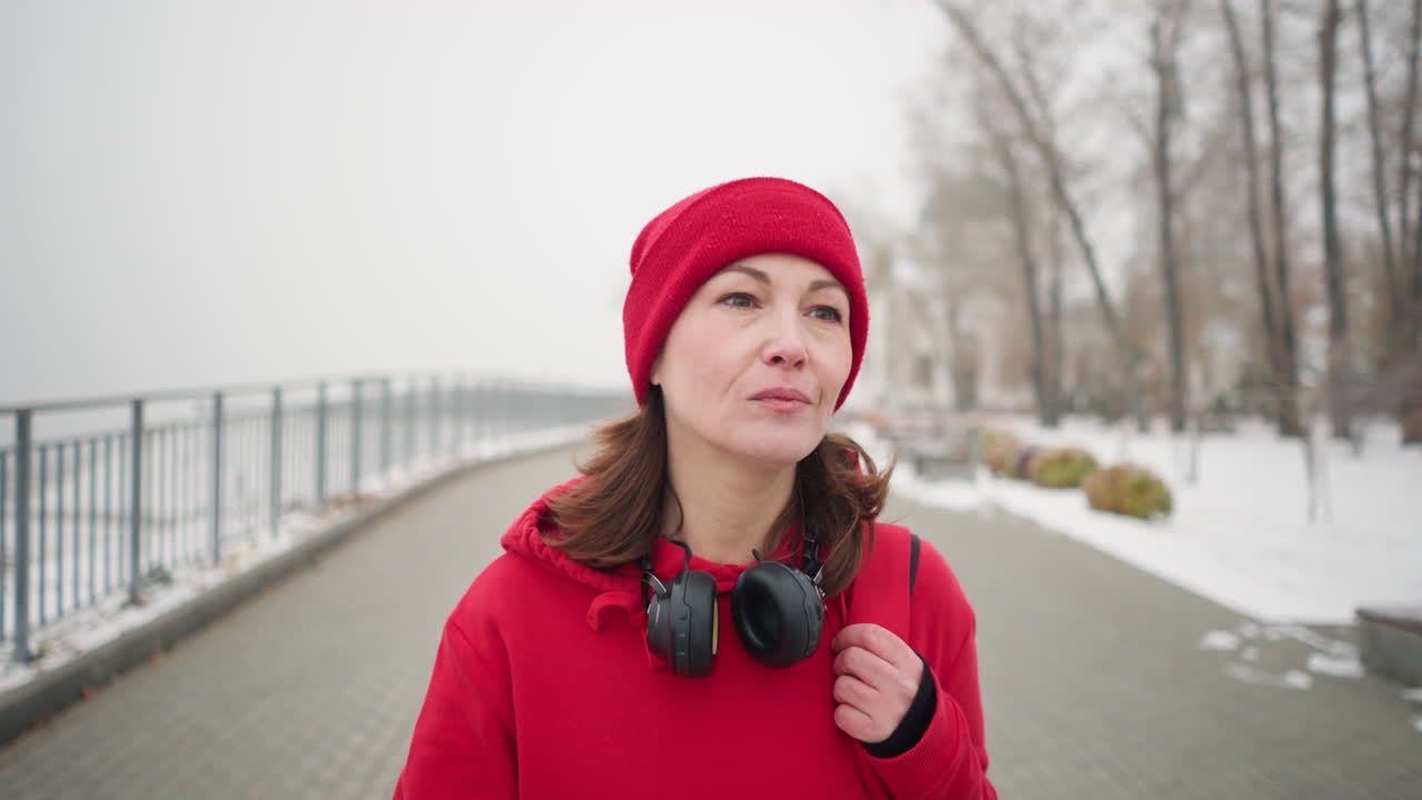 primer plano de una mujer con auriculares alrededor del cuello caminando por un camino entrelazado, sosteniendo la correa de la bolsa sobre el hombro con expresión tranquila, barandilla de hierro y árboles nevados en el fondo, en una atmósfera de niebla