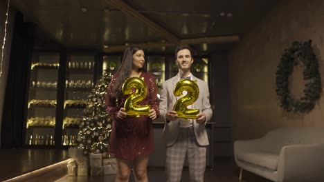 Woman and man wearing elegant clothes holding balloons with the numbers 22 while dancing and kissing each other in new year&#039;s party