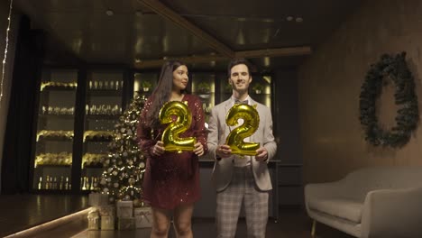 woman and man wearing elegant clothes holding balloons with the numbers 22 while dancing and kissing each other in new year's party