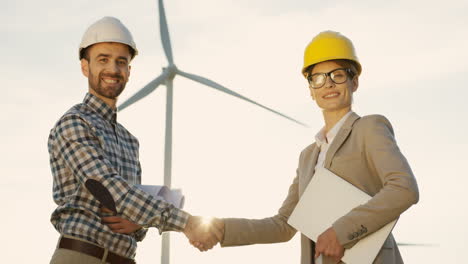 Vista-Inferior-De-La-Mujer-Caucásica-Y-Los-Ingenieros-Del-Hombre-Que-Usan-Un-Casco-Que-Se-Dan-La-Mano-Y-Miran-La-Cámara-En-La-Estación-Eólica-De-Energía-Renovable