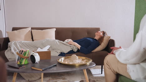 healing seeker in blue gown reclines on couch pulling blanket over herself while listening to therapist in calm therapy lounge with warm tones, soft lighting, and comforting atmosphere for healing
