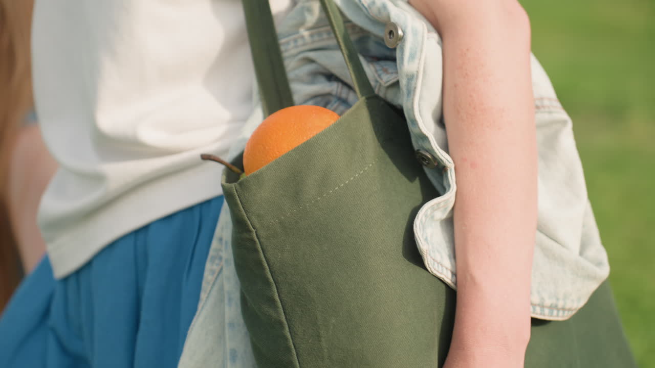 hand view of lady walking outdoors carrying orange peeking black canvas bag beside denim jacket on shoulder sunlight illuminating fruit and fabric textures casual style vibrant summer scene