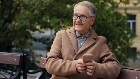 Anciano-Con-Gafas-Y-Abrigo-Sentado-En-El-Banco-Y-Enviando-Mensajes-De-Texto-Mientras-Toca-El-Teléfono-Inteligente-En-El-Parque-En-Otoño