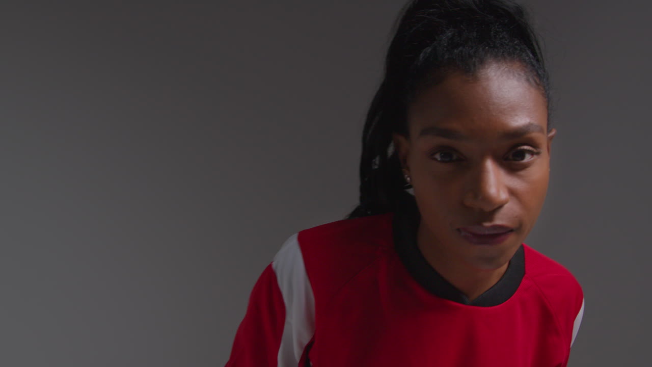 Studio Portrait Of Serious Female Football Or Soccer Player Wearing Team Shirt Holding Ball Under Her Arm Shot Against Grey Background 3
