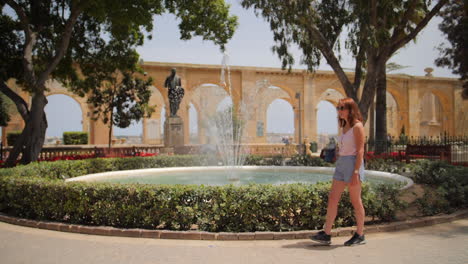 joven mujer con estilo caminando a lo largo de la fuente de manantial de agua mientras el agua salpica en cámara lenta en los jardines de la barraca superior en valletta, malta