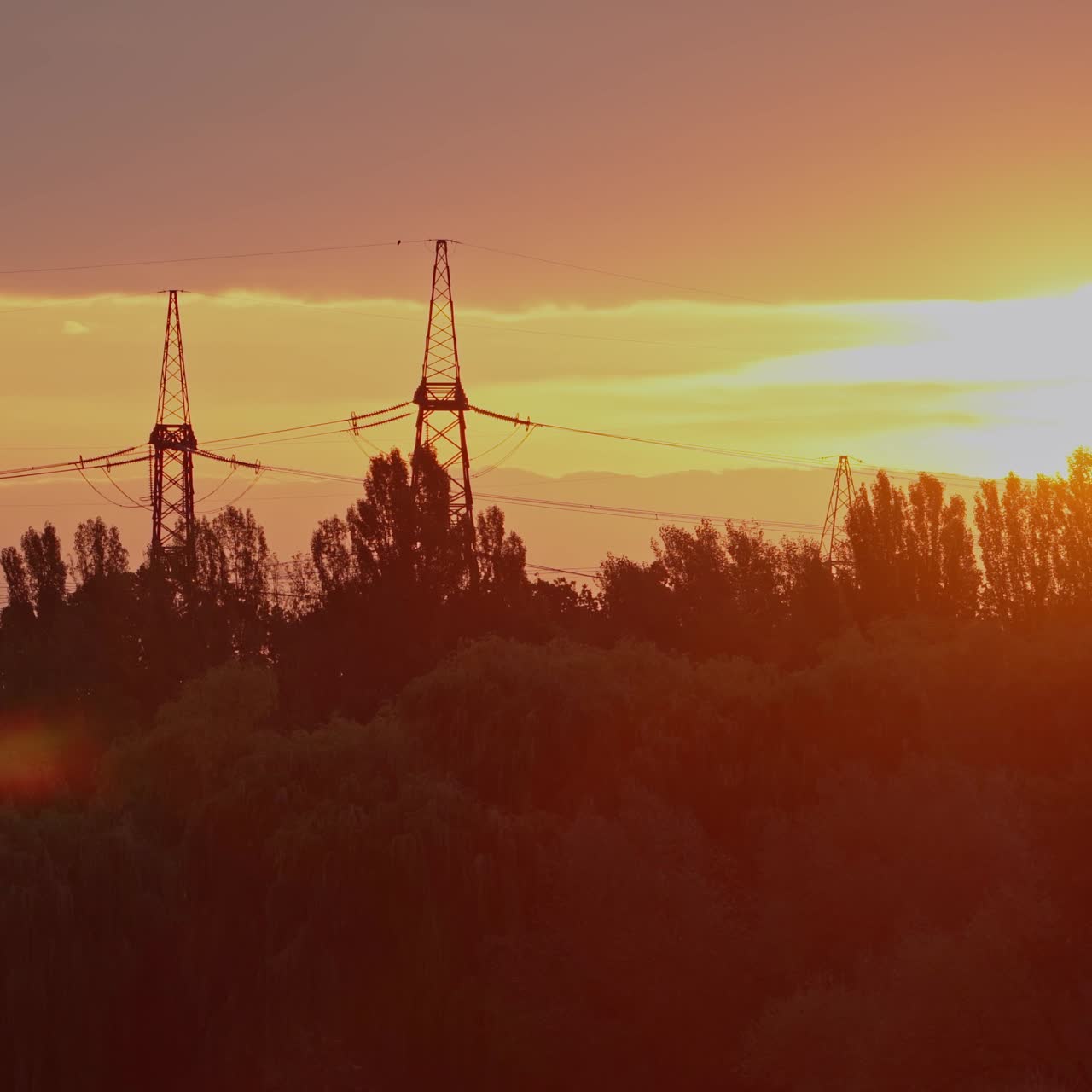 Tall electricity pylons at sunset. High voltage transmission towers and electric lines on the golden light. Panoramic view of sunset over the trees and electric towers in the evening.
