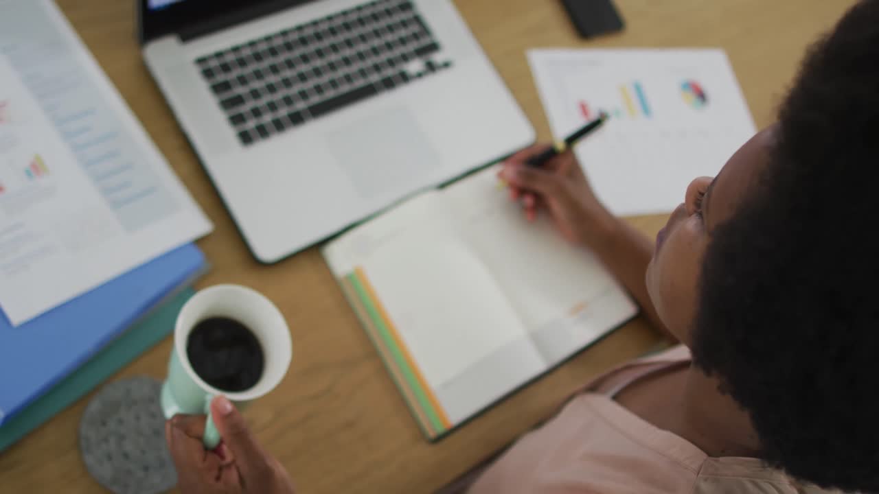 Happy african american woman sitting at table using laptop, drinking coffee