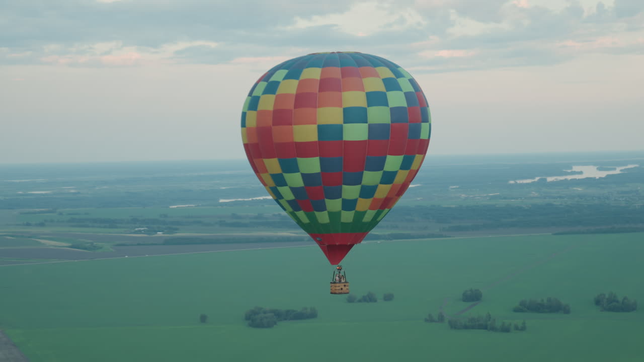 distant colorful hot air balloon drifting over expansive farmland with winding river on horizon against soft evening sky offering tranquil aerial scene of rural landscape