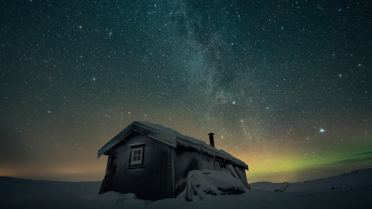 Small cabin under the milky way and aurora borealis. Cold winter night timelapse.
