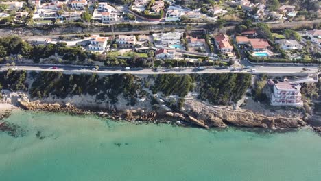 drone-flies-along-a-beautiful-coast-with-a-beach-and-a-street-with-some-traffic,-nice-houses