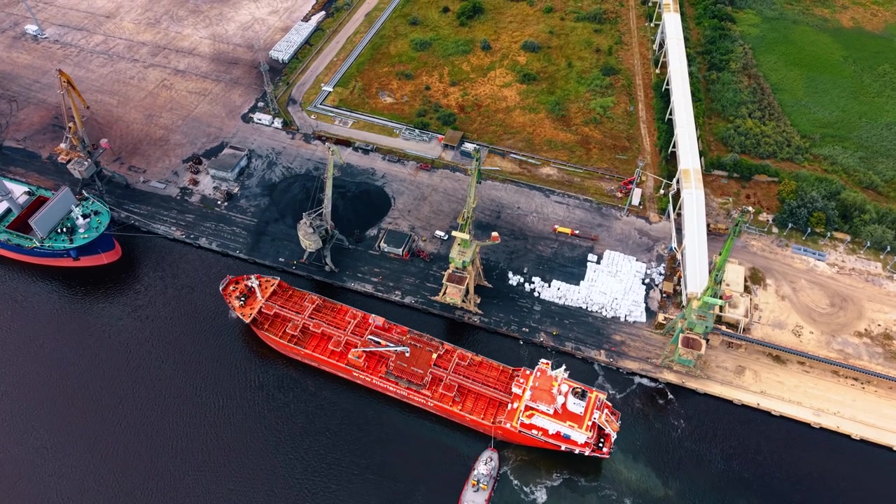 Varna, Bulgaria, 16 giugno 2026: Petroliera rossa che passa gru e cumuli di stoccaggio alla rinfusa. Vista verticale cattura un'imbarcazione accanto alla banchina, sacchi di carico bianchi, scorte scure e nastri trasportatori elevati