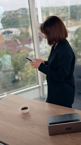 woman stands leaned over her laptop. lady in black jacket takes her phone and goes to the window. vertical video.