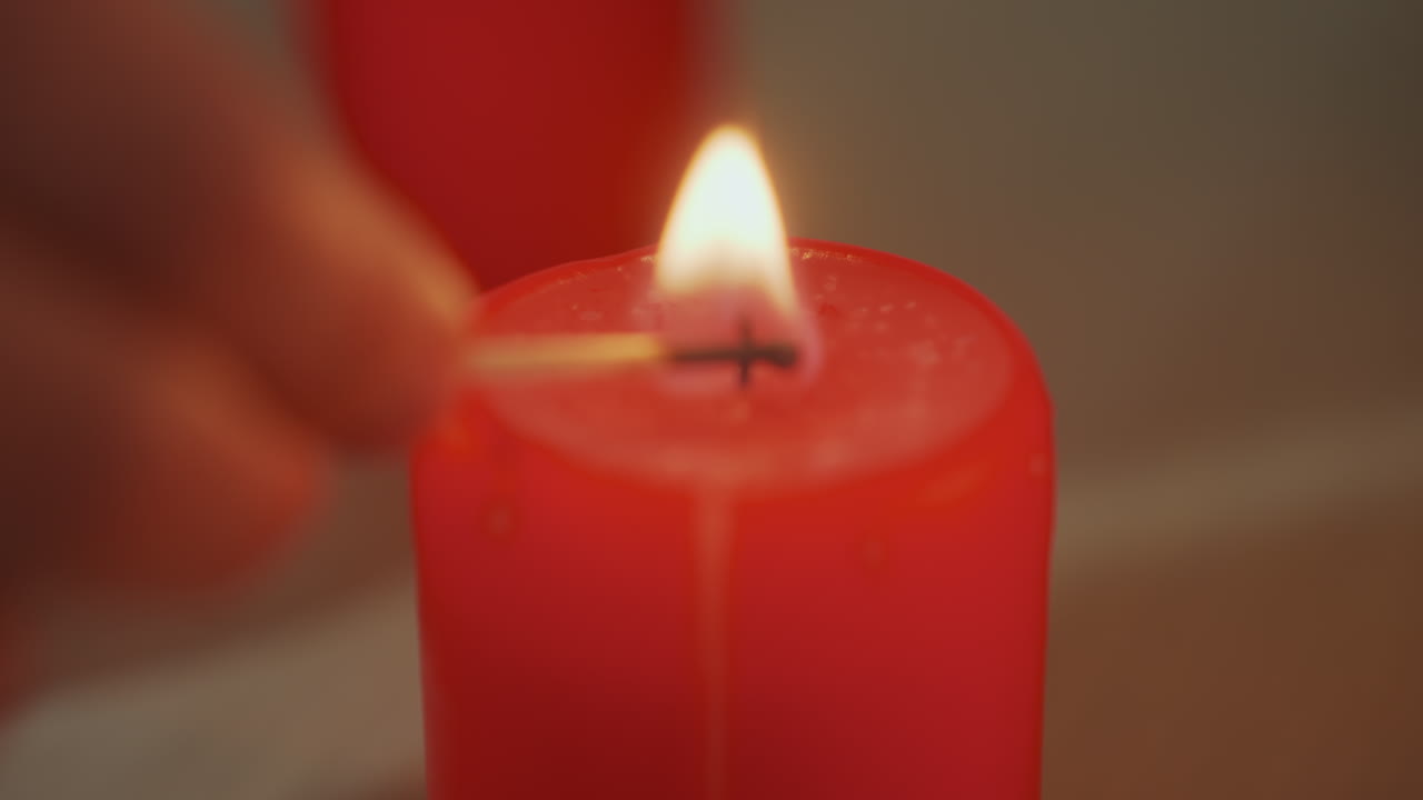 Candela rossa accesa a mano, primo piano: fiammifero accende lo stoppino e crea un bagliore caldo, goccioline di cera che si sciolgono, accogliente atmosfera serale da spa perfetta per massaggi e meditazione, stato d'animo rilassante e luce ambrata soffusa.