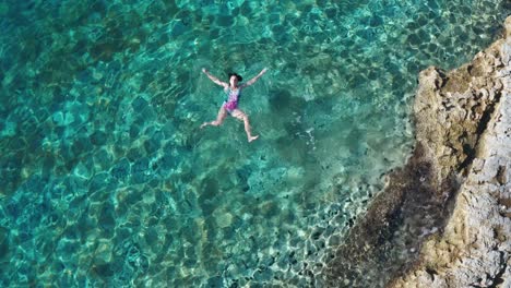 4k drone footage woman floating on her back near rocky coast on sea surface in transparent turquoise water