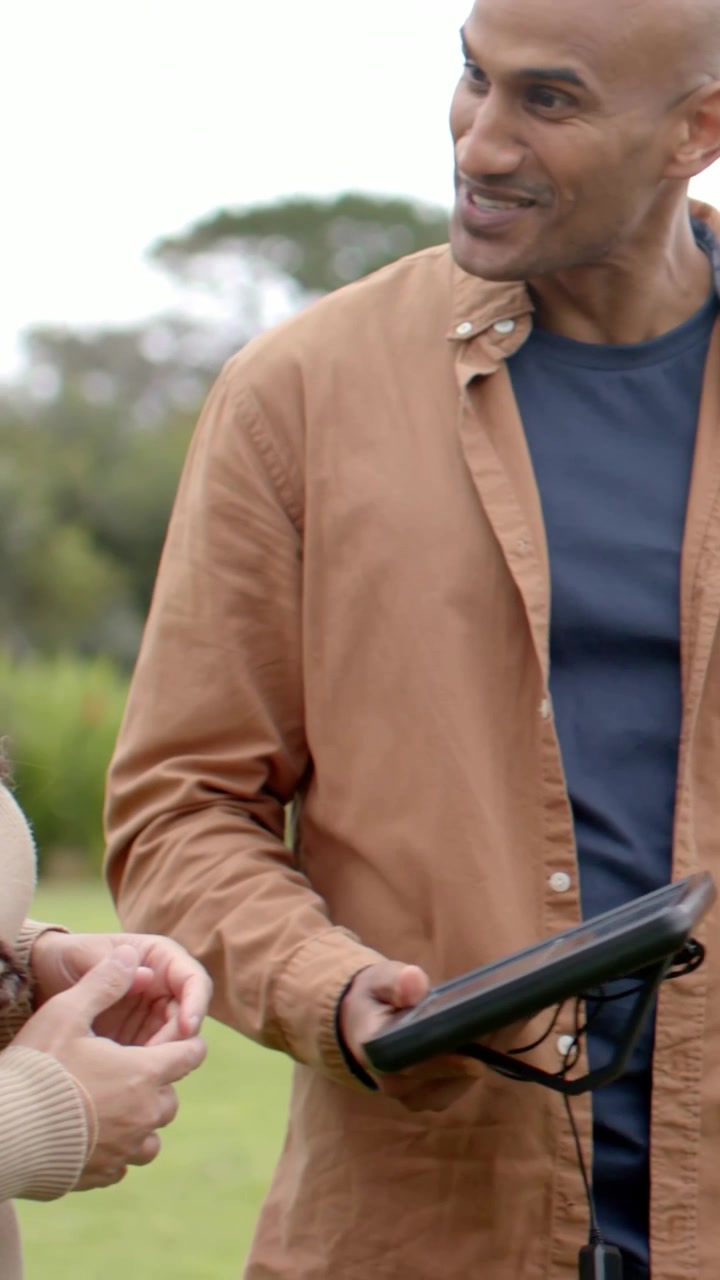Video verticale: Donna in maglione che parla al parco, guarda un uomo che si avvicina e mostra un tablet, con spazio per il testo.