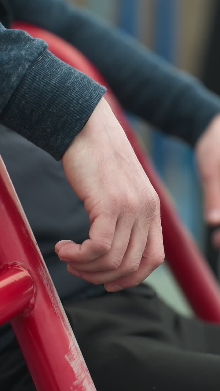 Close-up of a person's hands resting on a workout equipment as he shakes his leg, with a blurred view of a boy playing football and blue poles in the background
